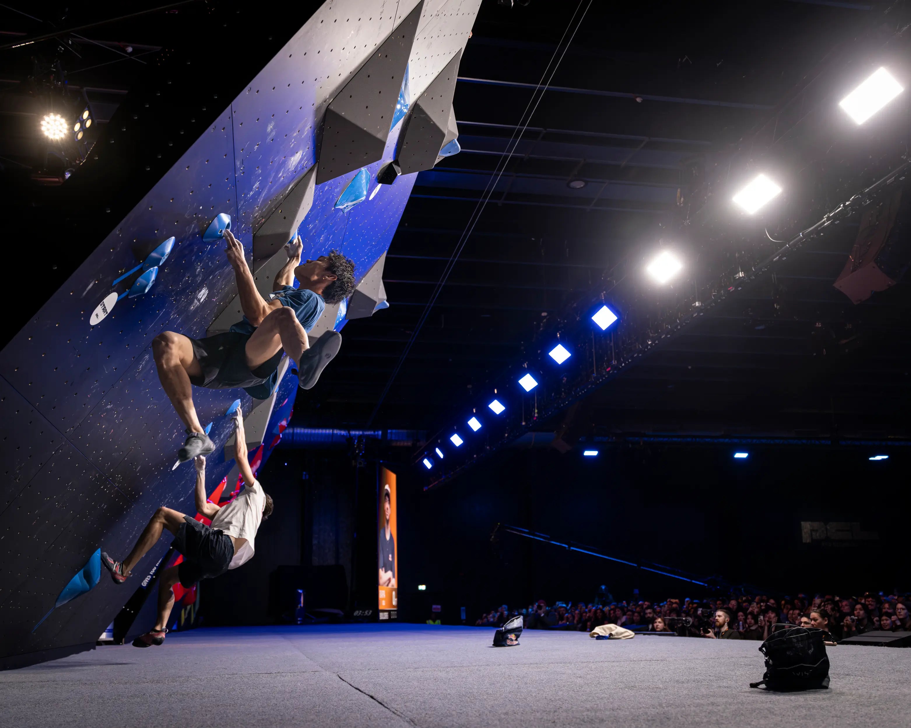 Person climbing a rock wall in an indoor climbing gym with spectators watching.